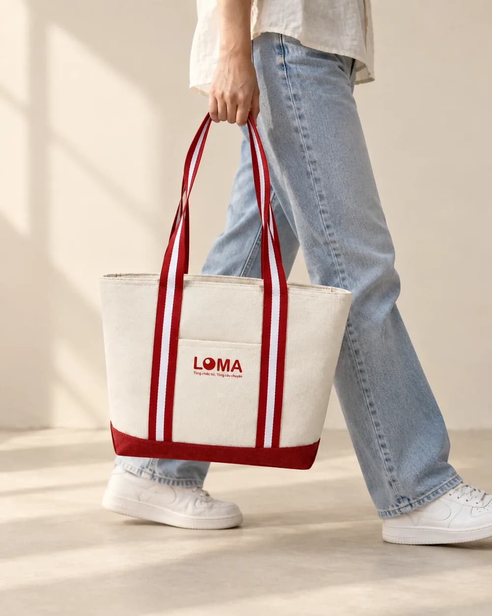Tay xách túi tote canvas quai sọc đỏ khi bước đi, quần jeans xanh nhạt, logo LOMA ở mặt trước