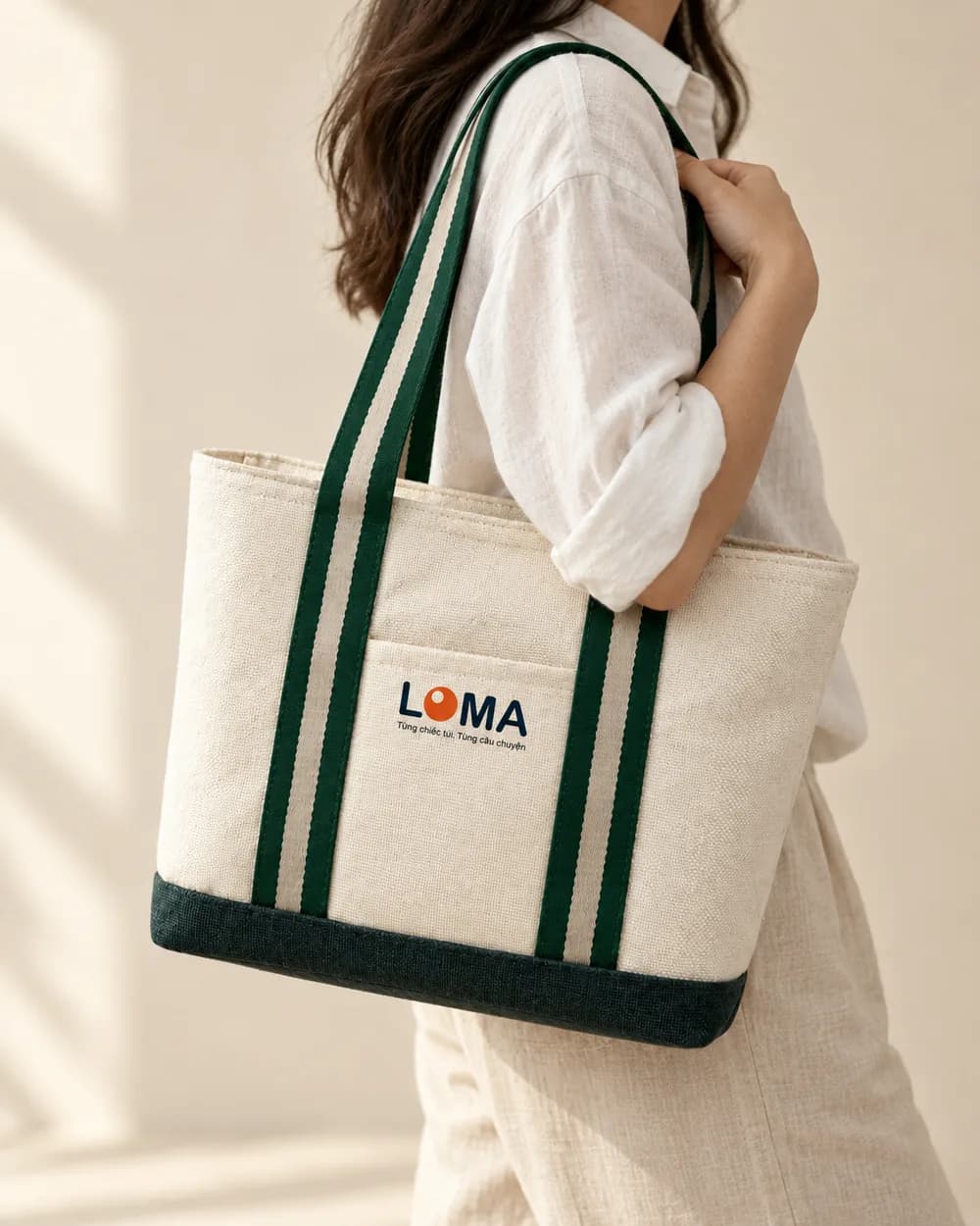 Túi tote canvas quai sọc xanh rêu khoác trên vai, áo sơ mi linen trắng, logo LOMA ở mặt trước